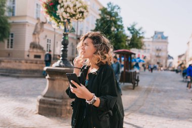 Romantic adorable happy girl with puffy hair wearing black jacket walking in city with smartphone in sunny warm day. Stylish girl exploring city in spring day.