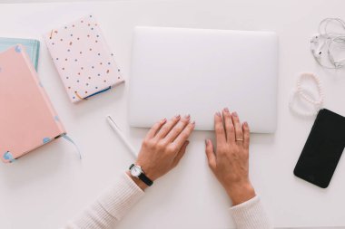 Top view of woman's hands typing on laptop. Work desk with smartphone, laptop, notebooks and headphones. Top view of woman sitting behind desk and making notes in documents.