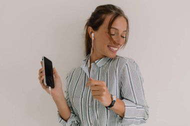Excited lovely goo-looking woman with dark hair wearing striped shirt dancing, having fun and listening to music with closed eyes and lovely smile against while wall.