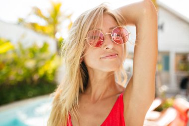 Portrait of attractive happy smiling blonde girl with long hair posing on exotic resort. She wears ts-hirt and round pink glasses 