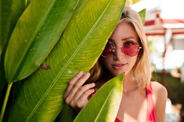Close up portrait of lovely cute blond girl in round pink glasses covering face by leaf and smiling to camera in exotic garden