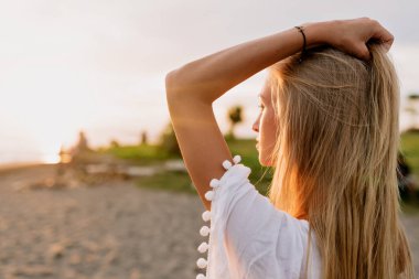 Close up portrait from back of blond long-haired woman looking straight to the ocean in sunlight on sandy beach. Rest and recovery concept. High quality photo