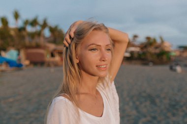 Romantic cute lovely girl with blue eyes and long blond hair looking straight to the camera on sandy beach near the ocean in sunlight 