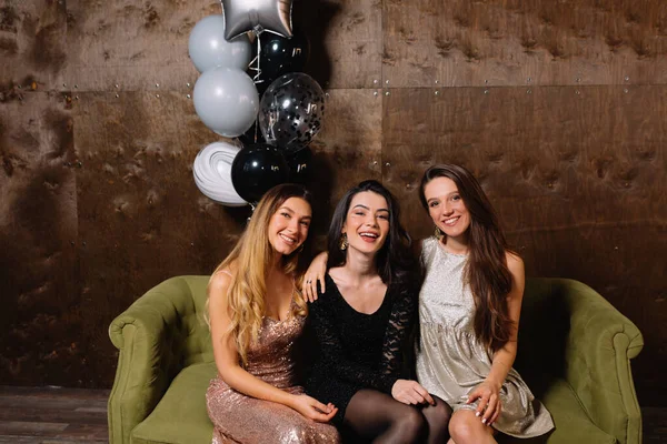 Smiling happy girl in festive dresses posing to camera with balloons over dark background. Close-up photo of refined women with stylish makeup relaxing at birthday party. Portrait of chilling girls