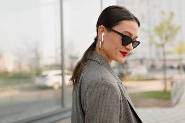 close UP PORTRAIT OF CHARMING ELEGANT WOMAN WITH DARK HAIR AND RED LIPS WEARING DARK SUNGLASSES AND GREY JACKET LOOKING DOWN WITH WONDERFUL SMILE AGAINST MODERN BUILDING . 