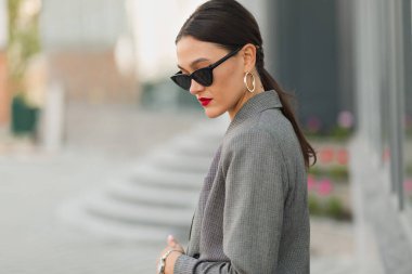 Attractive girl in grey jacket and dark glasses wearing golden earrings posing outside. Dark-haired woman with handbag and glasses looks away in city