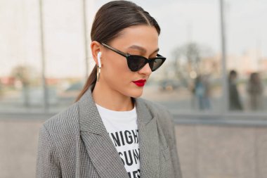 Elegant young woman with red lips and dark hair wearing grey jacket and dark sunglasses in headphones smiling and looking away outdoor