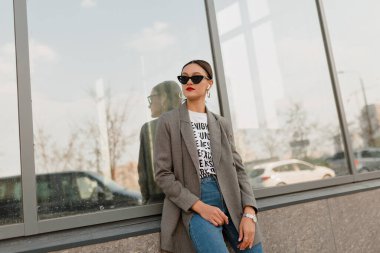 Appealing stylish woman with dark hair and red lips wearing grey jacket and jeans posing to camera against modern glass building in warm day