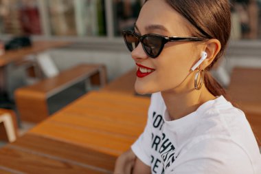 Close up profile portrait of smiling pretty girl with wonderful smile wearing dark sunglasses with red lipstick is listening music in wireless headphones and resting outdoor in sunlight 