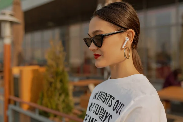 Trendy woman with dark hair and red lips wearing dark sunglasses and white t-shirt is walking the city on sunset, enjoying music and having fun outdoor.