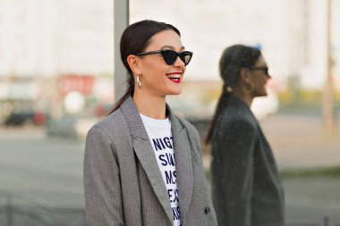 Smiling happy exited woman with wonderful smile and red lips wearing dark glasses, golden earrings, grey jacket walking iin city in business district with happy emotions