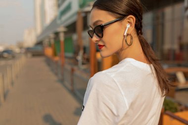 Close up portrait from back of adorable pretty brunette woman with red lips wearing dark sunglasses and golden earrings posing to camera , enjoying sunset in the city, resting during the walk.