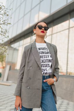 Confident young light-skinned woman walking along city street. Brunette in grey jacket and white t-shirt and jeans. Emotions and states of mind, concept.