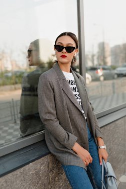 Trendy girl with dark hair in grey jacket and jeans in dark sunglasses looking aside and posing against modern city building. Stylish woman in eyeyglases sitting outdoors