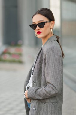 Dark-haired woman with red lips wearing dark glasses in grey jacket and white t-shirt walks outside. European lady smiles and moves outdoors.