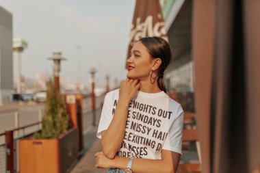 Outdoor close up portrait of attractive pretty brunette woman with collected hair and red lips posing outdoor in sunlight on blue city background 