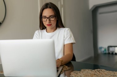 Photo of brunette woman look on laptop at home. Attractive woman smiling in glasses working at home with laptop on the table. Concept of freelance lifestyle