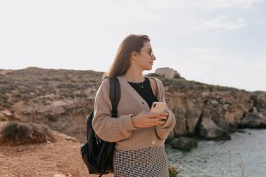 Profile portrait of adorable cute lovely girl with loose hair wraith knitted jacket and shirt with backpack looking to the ocean with happy smile in sunlight on background of cliffs 