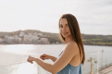 Close up portrait of romantic cute lady with loose dark hair wearing blue t-shirt is posing to camera on balcony in sunlight against lake