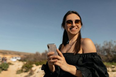 Smiling happy successful woman with snow-white smile in sunglasses is holding smartphone and looking aside against blue sky in sunny day 
