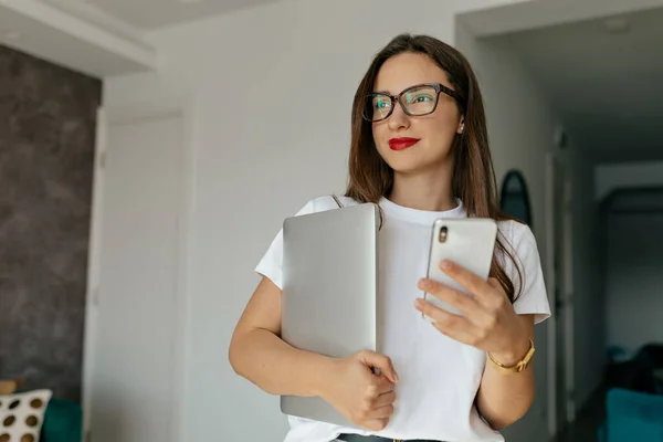 Independent pretty woman with dark hair, red lips in eyeglasses is holding laptop and smartphone and looking at window while working from home