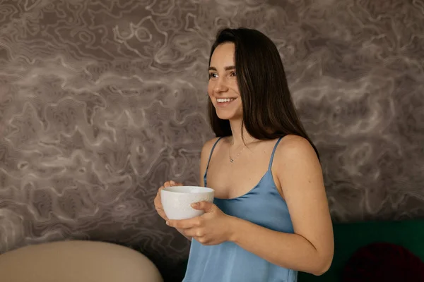 Happy woman with wonderful smile and dark loose hair wearing blue home suit is drinking coffee after morning wake up. Charming girl is making morning rituals at home