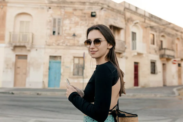 Happy charming caucasian woman with happy smile looks at camera relaxing on street. Brunette in sunglasses wears spring top and skirt. Lifestyle concept
