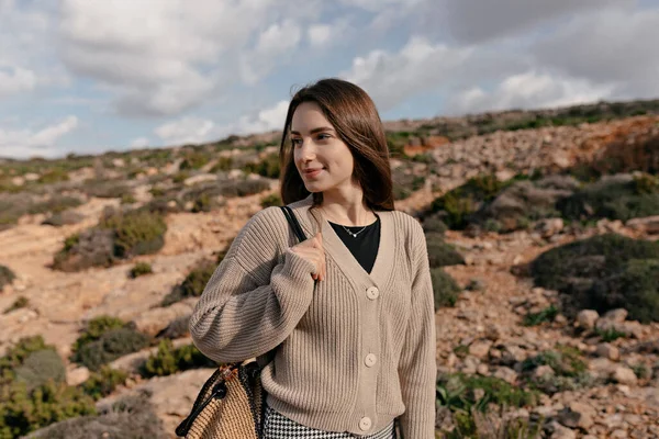 Lovely cute charming woman with dark loose hair wearing knitted shirt and holding bag is looking aside with happy smile against cliffs in sunlight