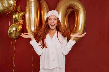 Çekici beyaz kadın, izole edilmiş arka planda kameraya poz veren ağzı açık bir kadın görünce şaşırıyor. Pembe pijamalı, kızıl saçlı, çekici kadın, el üstünde tutulur. 