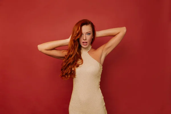 Stock image Amazing stylish European woman with long wavy red hair wearing golden dress is posing to camera with raised up hands over isolated red background