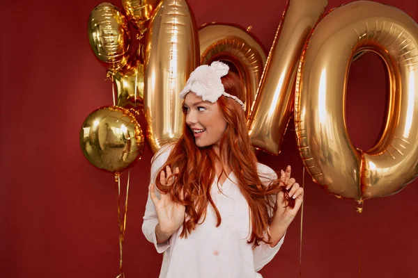 Close up profile portrait of adorable charming girl with wavy red hair wearing sleeping mask on the head and pink pajama is looking aside and smiling over red background with golden balloons