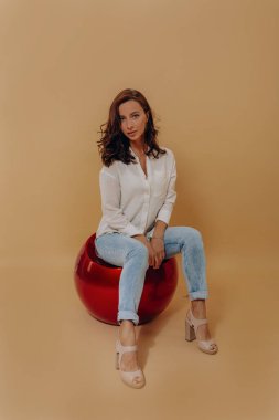 Beyaz gömlek, kot pantolon ve topuklu ayakkabı giyen, bej arka planda fotoğraf çekiminin keyfini çıkaran, hoş, koyu renk saçlı bir kadının tam boy portresi. 
