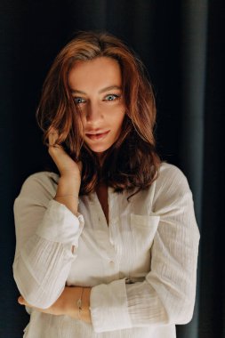 Büyük mavi gözlü, koyu renk dalgalı saçlı, koyu arkaplanda gülümseyerek kameraya poz veren çekici ve şık bir kadının portresini yakından çek.