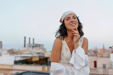 Çok güzel gülümseyen güzel bir kız, çenesine dokunuyor, kameraya gülümsüyor ve gün batımında şehir manzaralı bir terasta dinleniyor. 