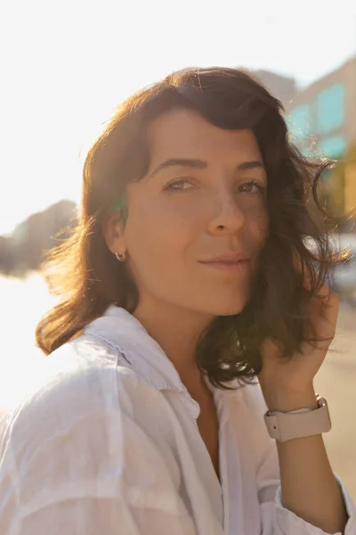 stock image Closeup portrait of pleasant girl with curly hair on street on sunset in the city. She wears white shirt. She is touching face and smiling to camera.