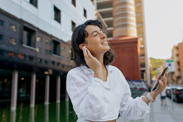 Relaxing calm lady with short hairstyle and gentle smile close eyes, listening to music and holding smartphone on city blurred background on sunset.