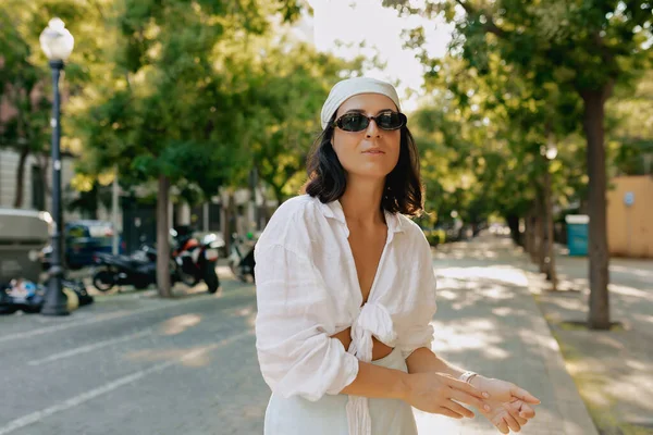 短い暗い髪型とヌードメイクとスタイリッシュな愛らしい女性は 太陽の光の下で街を歩く暗いサングラスで頭 白いシャツやスカートにショールを身に着けています — ストック写真