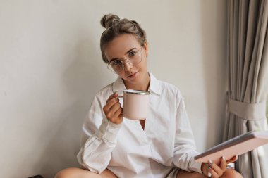 Saçları toplanmış ve çıplak makyajlı güzel bir kadın beyaz gömlek ve gözlük takıyor elinde bardak ve defter tutuyor ve izole bir arkaplanda kameraya bakıyor. 