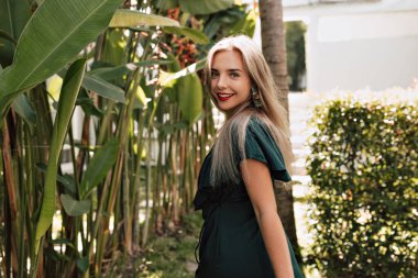 Kırmızı dudaklı ve uzun açık renk saçlı çekici bir kadın, güneşli bir günde, egzotik kısımların arka planında mutlu bir gülümsemeyle omzunun üzerinden bakıyor. 