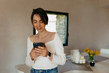 Kotlu, beyaz gömlekli, siyah saçlı, romantik ve çekici bir bayan, evde güneş ışığında akıllı telefon kullanırken gülümsüyor. 