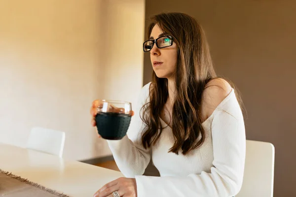 Incredible cute lady with loose hair wearing glasses and white blouse is enjoying morning coffee and looking at window. Concept lifestyle, using technology.