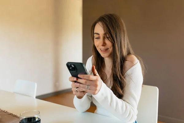 Image of beautiful woman smiling at phone . Cheerful caucasian young woman chatting online on smartphone in white blouse on home. Concept of use technology
