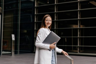 Kameraya gülümseyen, güzel görünümlü, mutlu bir şekilde poz veren, beyaz ceketli ve kot pantolonlu biri şehirde dizüstü bilgisayar tutuyor..