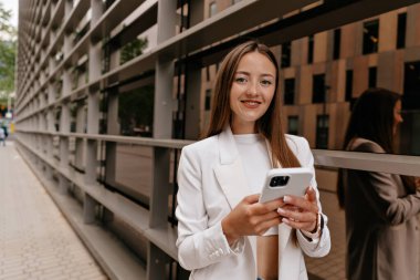 Elinde telefonla kameraya bakan neşeli, tapılası bir kadın resmi. Şehirde akıllı telefonuyla dolaşan gülümseyen beyaz bir kız. Beyaz ceket giyiyor.