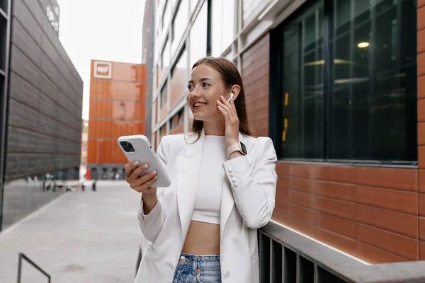 Profile portrait of adorable stylish woman with happy smile and looking aside while using smartphone and listening music while posing in business district in the city