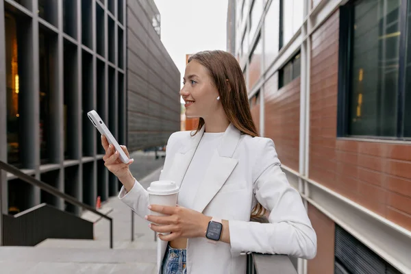 Profile outdoor portrait of pretty stylish girl in white jacket and top is using smartphone and drinking coffee in city. Young lady staying near to wall with phone. Street style concept.