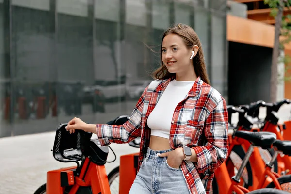 European stylish pretty woman with happy smile wearing white t-shirt and jeans in wireless headphones is looking straight with happy smile and posing on background red bikes in the city