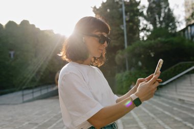 Kısa dalgalı saç stili olan şık, sevimli bayan beyaz tişört giyiyor ve siyah güneş gözlüğü akıllı telefon kullanıyor ve şehir parkında güneş ışığında dinleniyor. 