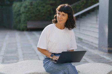 Kısa siyah saçlı, beyaz tişörtlü ve kot pantolonlu çekici bir kadın güneşli bir günde dizüstü bilgisayarda çalışıyor. Teknoloji kavramı 