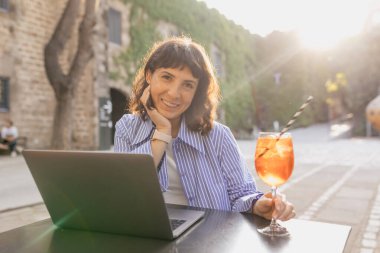 Kısa dalgalı saç stili, ıslak günlük kıyafetleri olan sevimli, sevimli bir kız şehirde güneşli bir parkta dizüstü bilgisayarla oturuyor ve elinde yaz kokteyli tutuyor. Şehir hayatı konsepti. 